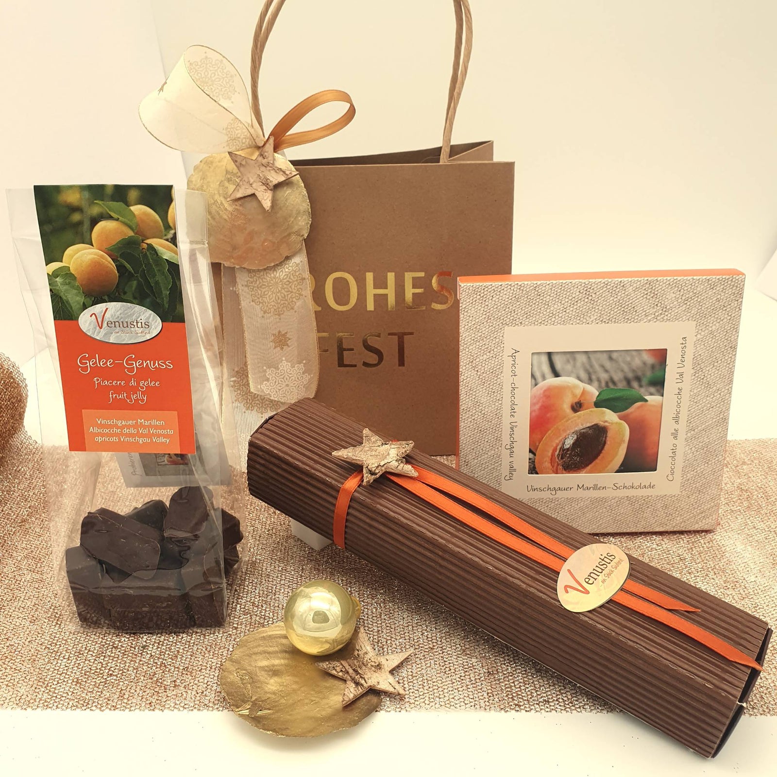 Weihnachtlich verpackte Venustis-Geschenke: Gelee-Genuss, Pralinenstange und Vinschgauer Marillen-Schokolade vor einer Tasche mit der Aufschrift Frohes Fest